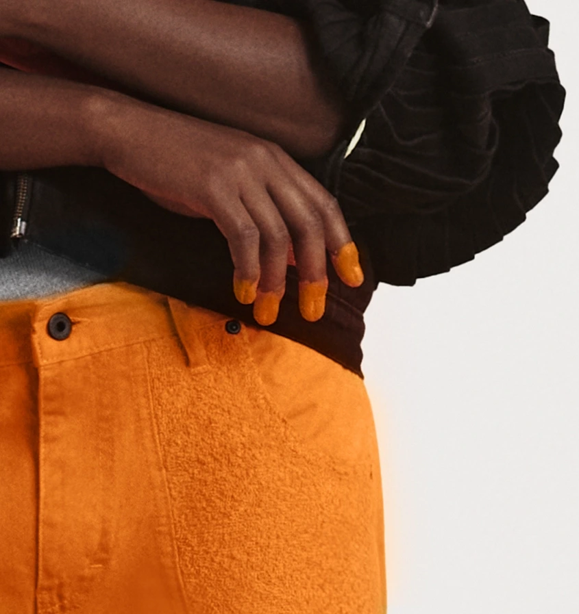 A close-up of a person wearing a black jacket and orange pants with a texture resembling Cheetos dust. Their fingers are covered in an orange residue, suggesting they have just eaten cheesy snacks. The image highlights the contrast between the dark tones of the jacket and the vibrant orange color of the pants and fingers.