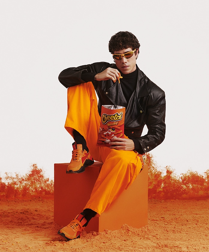 A close-up shot of the young man's hand resting on his bright orange pants, which feature a textured fabric resembling Cheeto dust residue. His fingers are visibly covered in the signature orange Cheetos seasoning, emphasizing the playful, messy eating experience. The leather sleeve of his jacket contrasts with the vibrant orange tones.